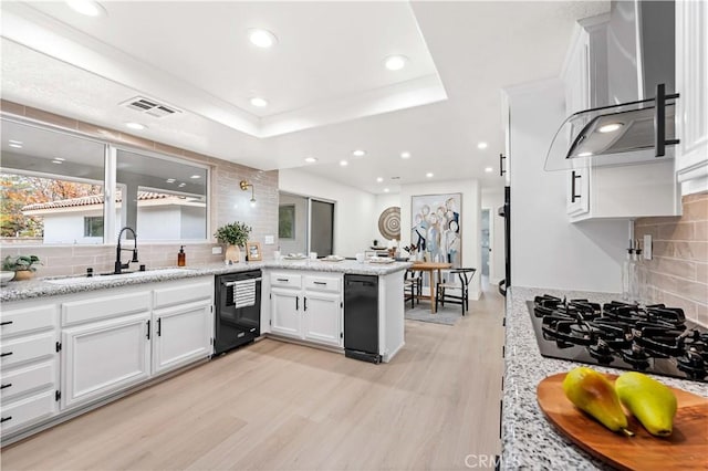 kitchen with a raised ceiling, a sink, black appliances, a peninsula, and wall chimney exhaust hood