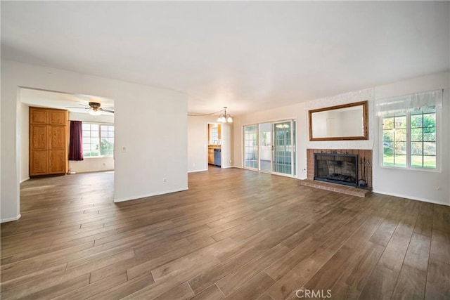 unfurnished living room with ceiling fan with notable chandelier, a brick fireplace, baseboards, and wood finished floors