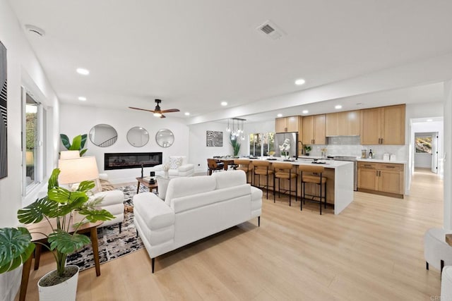 living area featuring light wood finished floors, recessed lighting, visible vents, a glass covered fireplace, and ceiling fan with notable chandelier