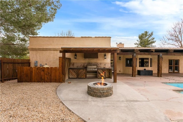 back of property featuring stucco siding, an outdoor kitchen, a patio area, fence, and a fire pit