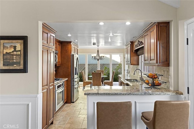 kitchen with a sink, light stone counters, stone tile flooring, stainless steel appliances, and an inviting chandelier