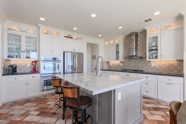 kitchen featuring visible vents, a center island with sink, a sink, stainless steel appliances, and wall chimney exhaust hood