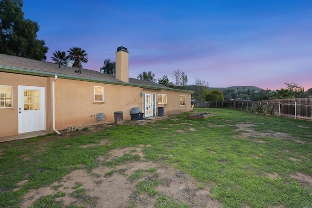 back of house at dusk featuring a yard, a fenced backyard, a chimney, and stucco siding