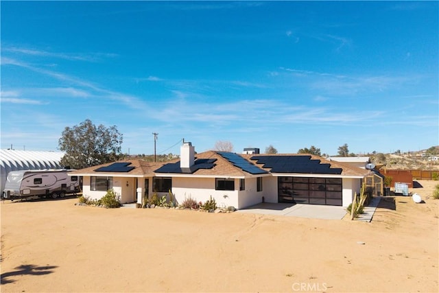 single story home featuring an attached garage, dirt driveway, roof mounted solar panels, a chimney, and stucco siding