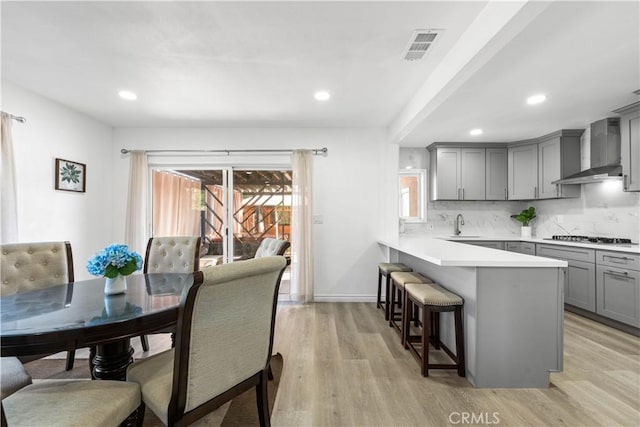 kitchen with a peninsula, a breakfast bar, light wood-style floors, gray cabinets, and wall chimney exhaust hood