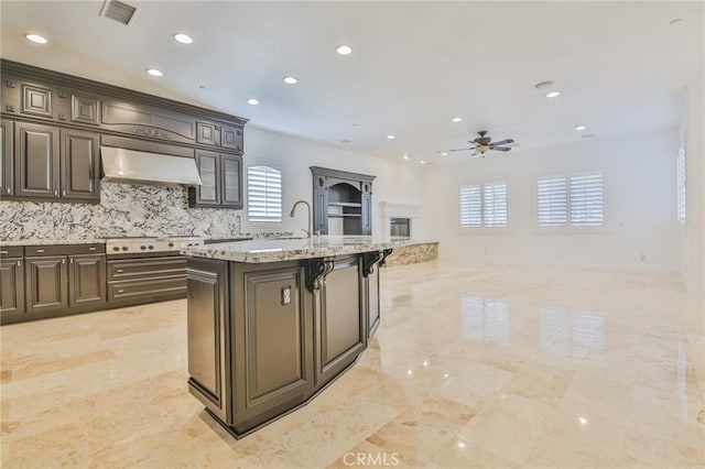 kitchen featuring light stone counters, visible vents, open floor plan, wall chimney range hood, and an island with sink