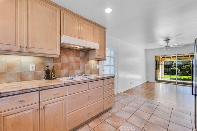 kitchen featuring tile counters, light brown cabinets, under cabinet range hood, and white stovetop