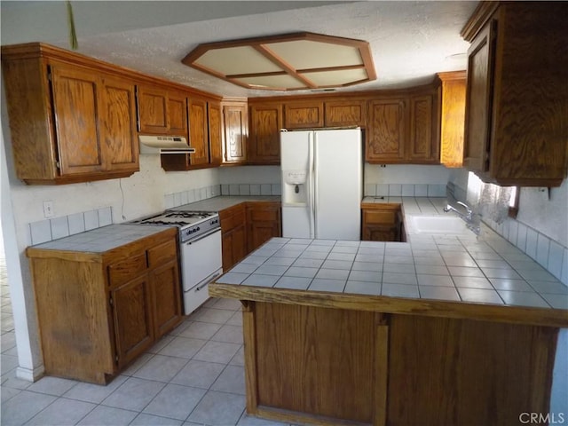 kitchen with white appliances, a sink, tile counters, under cabinet range hood, and brown cabinets