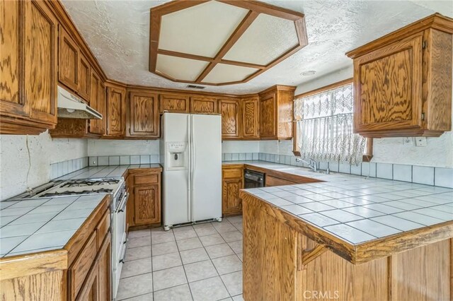11671 Jacaranda Ave, Hesperia, CA 92345 - kitchen with white appliances, tile counters, brown cabinetry, a peninsula, and under cabinet range hood