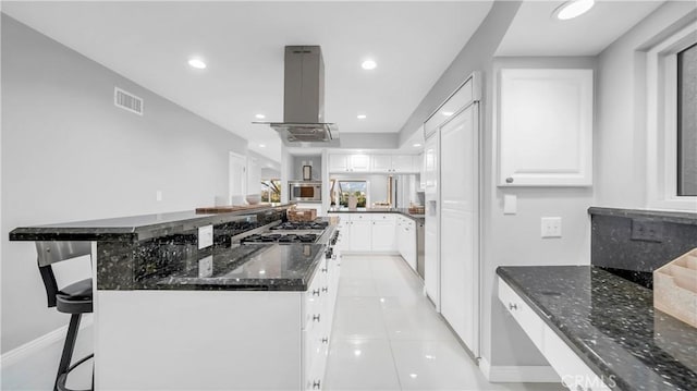 kitchen with light tile patterned floors, visible vents, stainless steel microwave, island exhaust hood, and white cabinetry