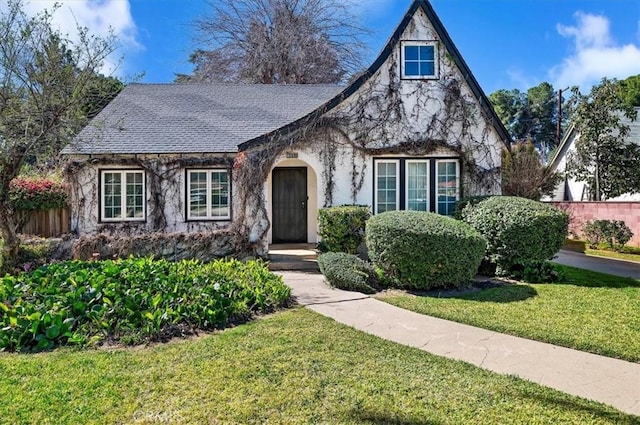 tudor house featuring a front yard, stone siding, roof with shingles, and fence