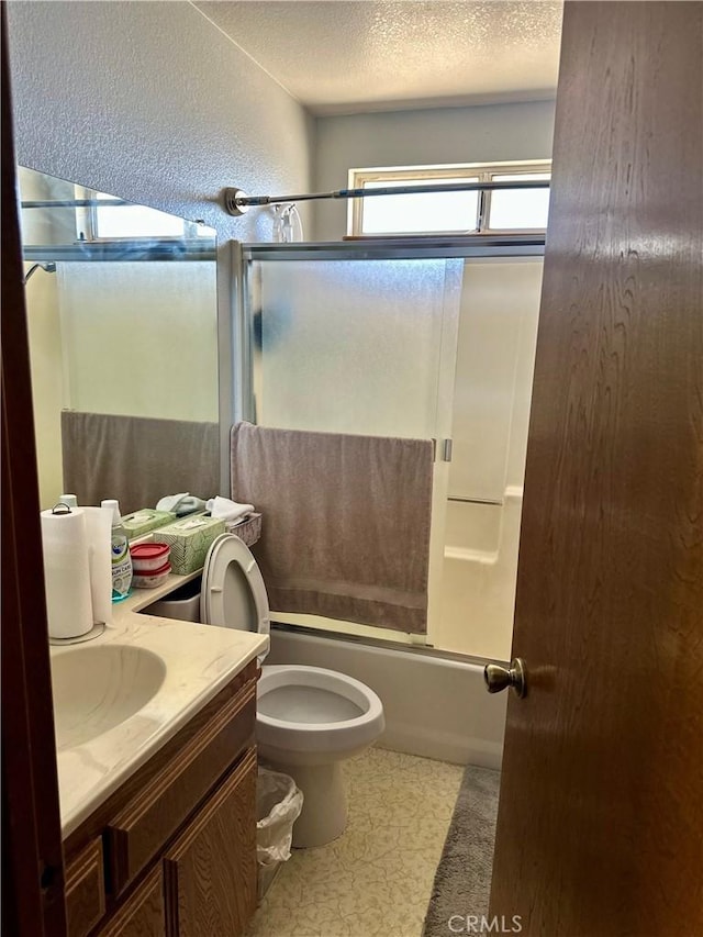 full bath featuring bath / shower combo with glass door, a textured wall, toilet, a textured ceiling, and vanity