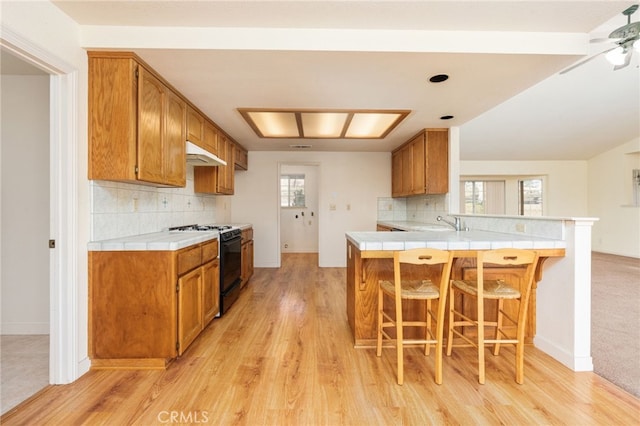 kitchen featuring black range with gas cooktop, tile counters, a breakfast bar, under cabinet range hood, and a sink