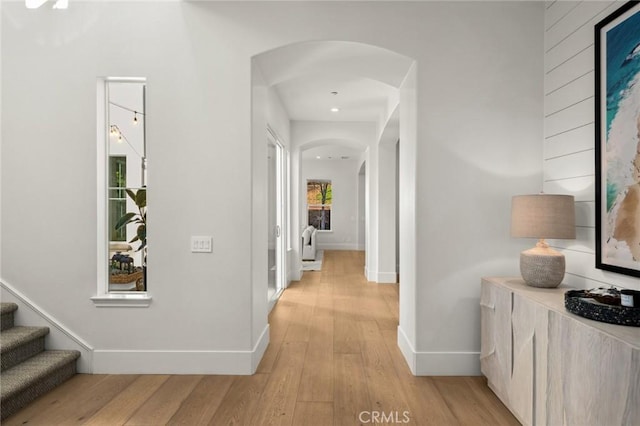 corridor featuring arched walkways, stairway, light wood-style flooring, and baseboards