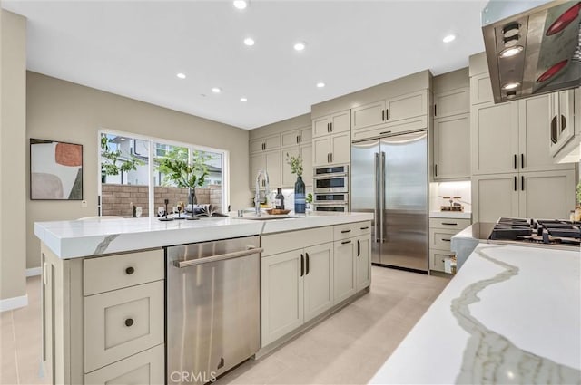 kitchen featuring light stone counters, a kitchen island with sink, ventilation hood, stainless steel appliances, and a sink