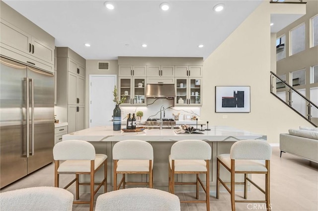 kitchen featuring tasteful backsplash, visible vents, glass insert cabinets, stainless steel built in refrigerator, and under cabinet range hood