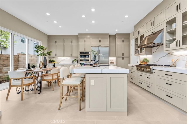 kitchen with an island with sink, appliances with stainless steel finishes, glass insert cabinets, under cabinet range hood, and backsplash