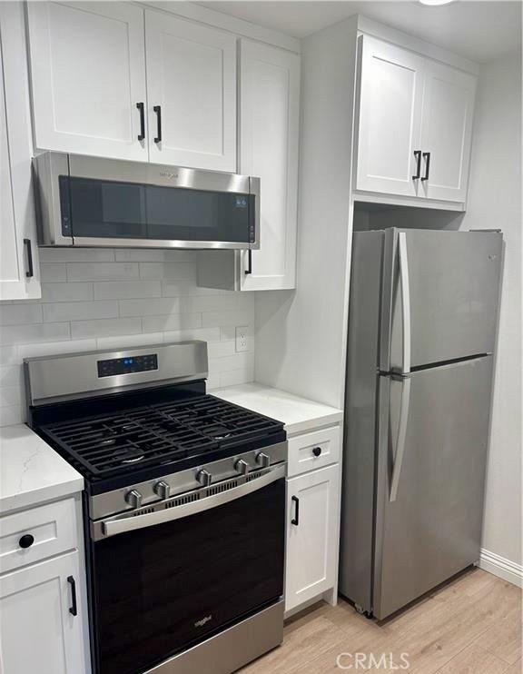 kitchen with light wood finished floors, appliances with stainless steel finishes, light stone counters, white cabinetry, and backsplash