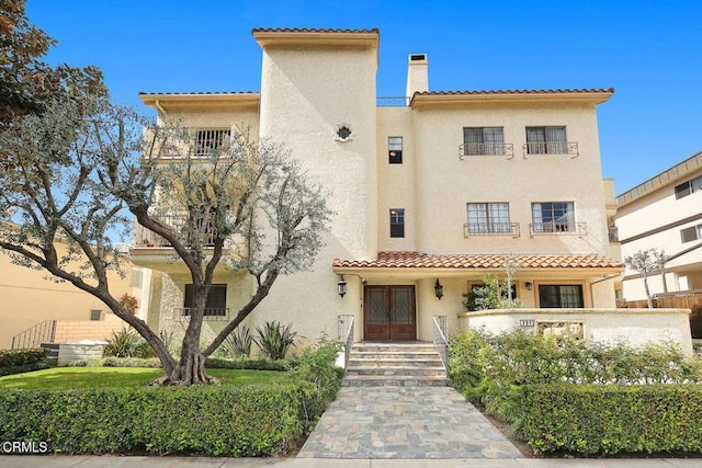 view of front facade featuring french doors, a chimney, a balcony, and stucco siding