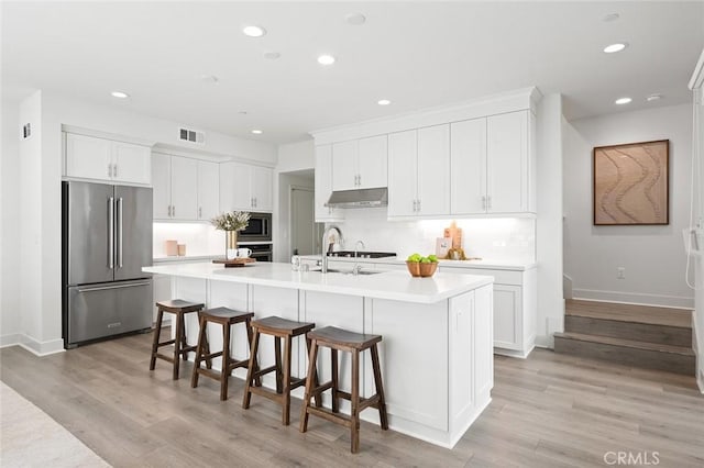 kitchen with under cabinet range hood, stainless steel appliances, white cabinets, light countertops, and a center island with sink