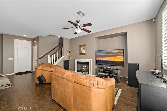 living room with dark wood-style floors, visible vents, a ceiling fan, a glass covered fireplace, and stairs