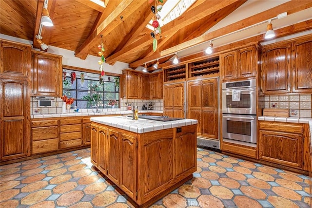 kitchen with a center island with sink, tile countertops, backsplash, lofted ceiling with beams, and double oven