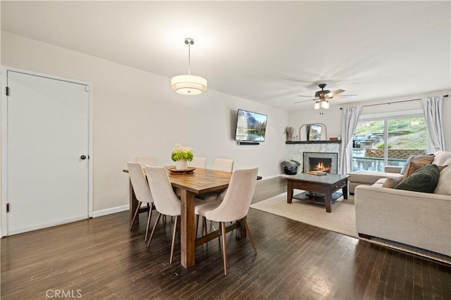 dining room with baseboards, dark wood-type flooring, a lit fireplace, and a ceiling fan