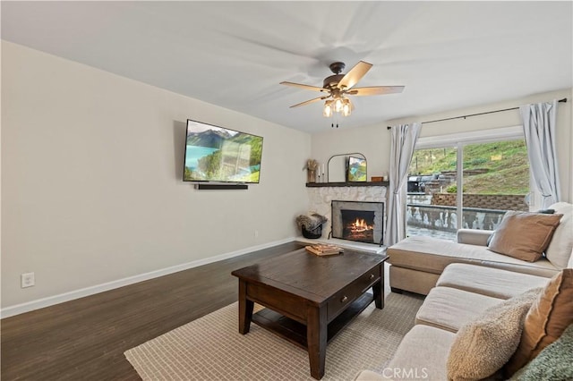 living room featuring ceiling fan, baseboards, a stone fireplace, and wood finished floors