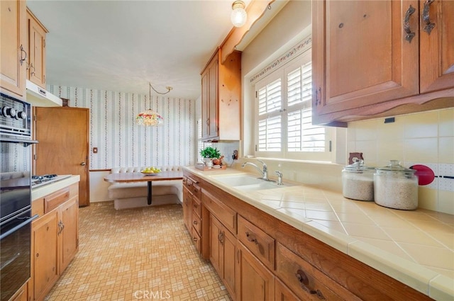 kitchen featuring brown cabinetry, wallpapered walls, a sink, tile counters, and under cabinet range hood