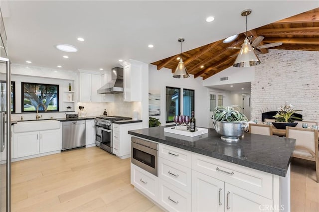 kitchen with lofted ceiling with beams, appliances with stainless steel finishes, wall chimney range hood, white cabinetry, and a sink