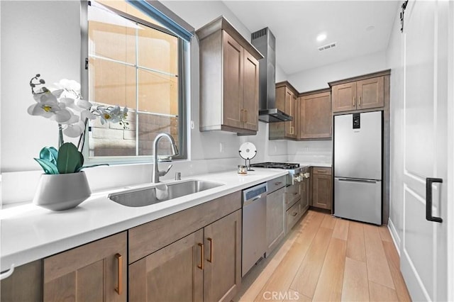 kitchen with light countertops, appliances with stainless steel finishes, light wood-style floors, a sink, and wall chimney range hood