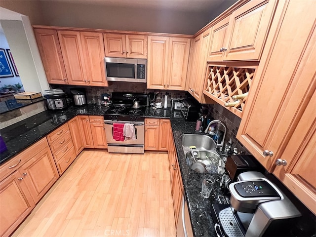 kitchen with appliances with stainless steel finishes, dark stone countertops, light brown cabinetry, light wood-type flooring, and a sink