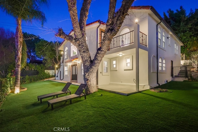 back of house at night featuring a patio, a yard, stucco siding, fence, and a balcony