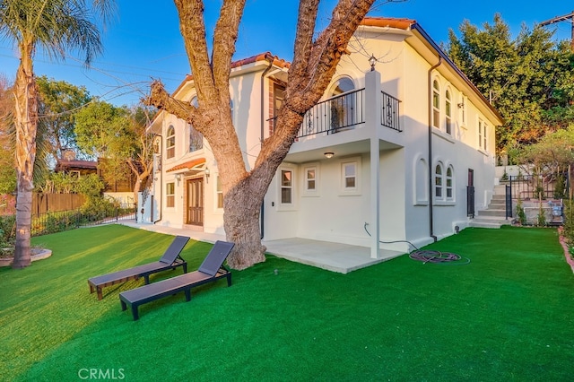 back of house with stucco siding, a lawn, a patio area, fence, and a balcony