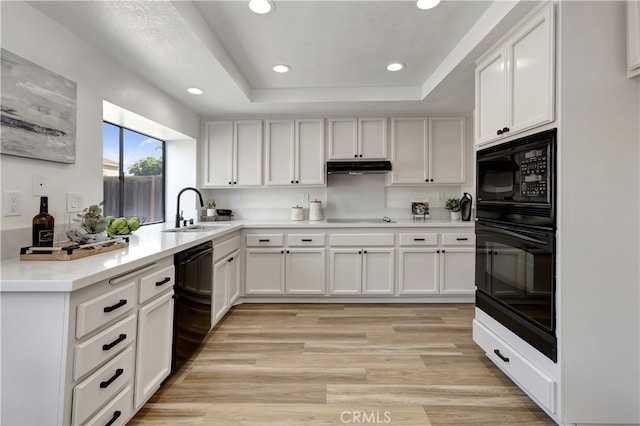 kitchen with black appliances, a sink, a tray ceiling, under cabinet range hood, and light wood finished floors