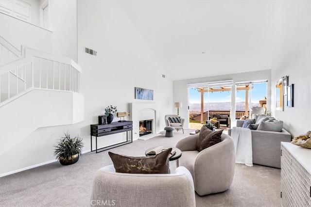 living room with visible vents, stairway, light colored carpet, a warm lit fireplace, and high vaulted ceiling