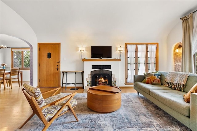 living area with a warm lit fireplace, arched walkways, wood finished floors, vaulted ceiling, and a notable chandelier