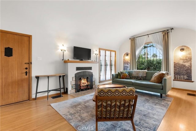 living area with visible vents, a fireplace with flush hearth, vaulted ceiling, wood finished floors, and baseboards