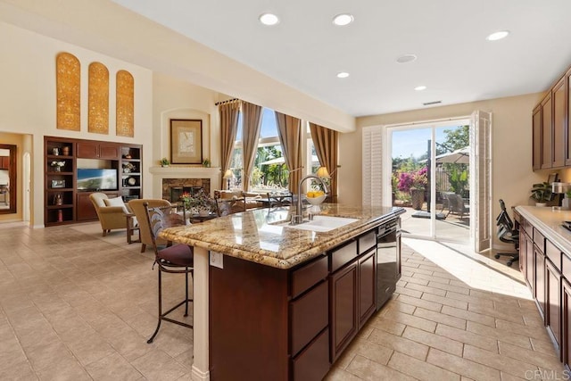 kitchen featuring dishwashing machine, a sink, a kitchen breakfast bar, a lit fireplace, and a center island with sink