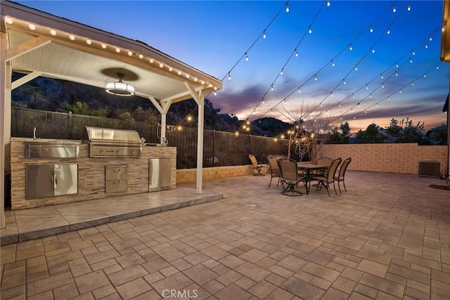 patio terrace at dusk featuring outdoor dining area, a fenced backyard, grilling area, and an outdoor kitchen