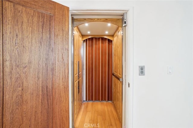 corridor featuring elevator, arched walkways, and light wood finished floors