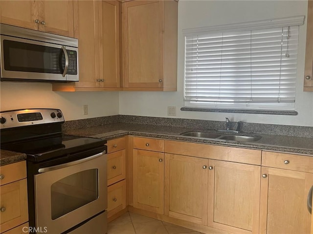 kitchen featuring light tile patterned floors, light brown cabinets, stainless steel appliances, a sink, and dark stone counters