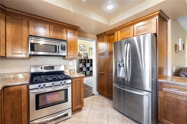 kitchen featuring brown cabinetry, light tile patterned floors, stainless steel appliances, and light stone countertops