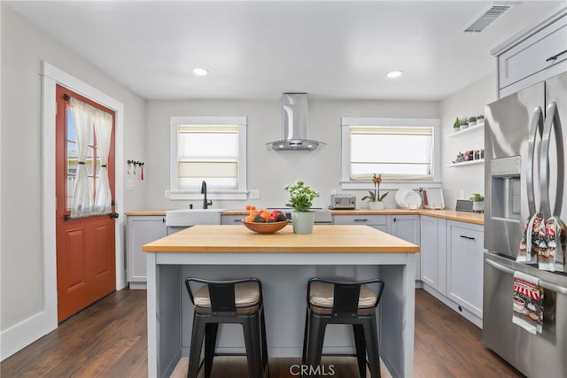 kitchen with stainless steel refrigerator with ice dispenser, a breakfast bar area, a sink, wood counters, and wall chimney range hood