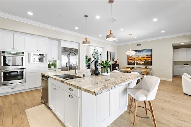 kitchen featuring stainless steel double oven, an island with sink, a sink, ornamental molding, and light wood-style floors