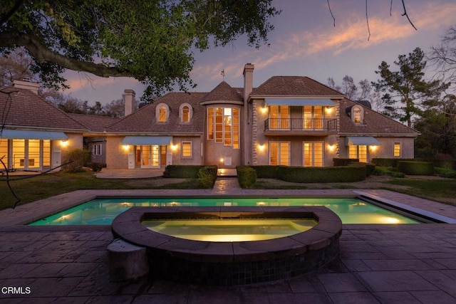 back of property at dusk with a balcony, a patio area, a chimney, and an outdoor pool