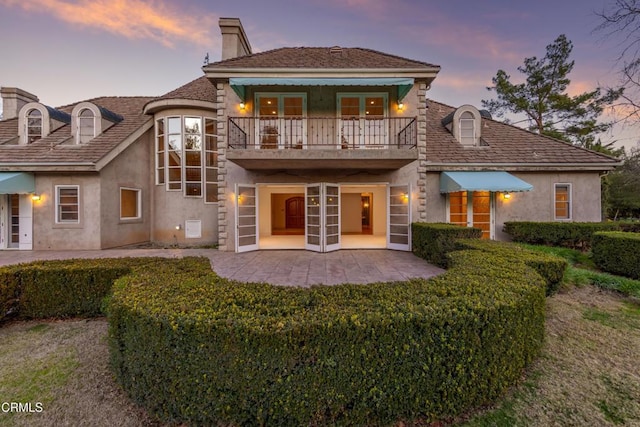 back of house at dusk with a chimney, a patio, a balcony, and stucco siding