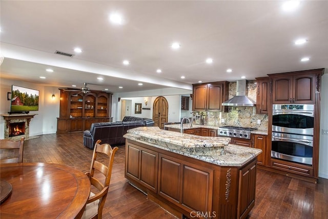 kitchen with a warm lit fireplace, dark wood-style flooring, visible vents, wall chimney range hood, and appliances with stainless steel finishes