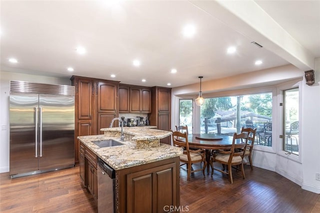 kitchen featuring dark wood-style floors, a center island with sink, stainless steel appliances, a sink, and light stone countertops