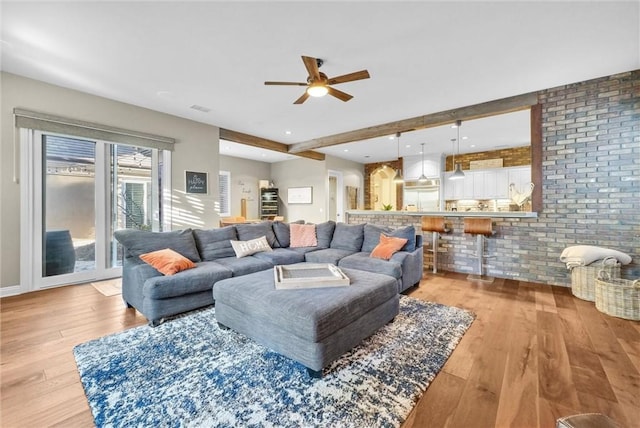 living area featuring ceiling fan, brick wall, light wood finished floors, and beamed ceiling